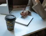 Kobieta notuje, zastanawiając się, czy okres próbny wlicza się do stażu pracy. Obok kubek kawy i tablet.