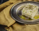 Ein Stück cremiger, vegetarischer Feta mit Kräutern und Olivenöl auf einem silbernen Teller, daneben ein rustikales Leinentuch.