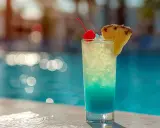 En blå och gul drink med körsbär och ananas står vid en pool. Perfekt för en solig dag.