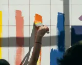 Różne rodzaje farb plastycznych ożywiają białą ścianę. Pędzel z tęczowym akcentem maluje pas pomarańczowy, obok żółty, koralowy i niebieski.