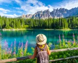 En kvinna med hatt och ryggsäck njuter av den vackra **Dolomiterna sommar**-utsikten över en turkos sjö, omgiven av skog och berg.