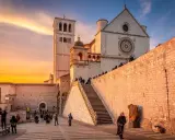 Assisi cosa vedere: la Basilica di San Francesco al tramonto, con la sua imponente facciata e la torre campanaria, illuminata da una calda luce dorata.