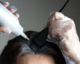 Hände mit Handschuhen tragen eine weiße Flüssigkeit auf dunkles Haar auf, um die Haarfarbe zu entfernen.