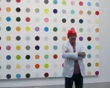 Damien Hirst steht vor seiner berühmten Punkt-Malerei, gekleidet in eine orangefarbene Mütze und einen weißen Blazer.