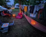 Una persona lee en una silla junto a una fogata, ropa secándose al aire y una hamaca iluminada. Disfrutando de una tranquila acampada al aire libre.