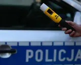 Policjant przeprowadza badanie trzeźwości nieletniego przy użyciu alkomatu. Na samochodzie widnieje napis "POLICJA".