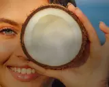 Donna sorride con un fiore tra i capelli, coprendo un occhio con una noce di cocco. Attenzione: olio di cocco sul viso fa male.