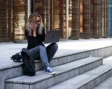 Kobieta z laptopem i telefonem siedzi na schodach. Jej styl przypomina nowoczesną **figure html**, pracującą zdalnie.