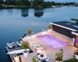 Lyxig pool med platssvetsad liner och belysning, omgiven av solstolar och växter. Båtar ligger vid bryggan.