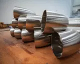 Pièces métalliques brillantes, probablement des coudes pour un poêle à bois conduit, empilées sur une surface en bois.