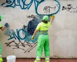 Dos trabajadores con uniformes verdes y amarillos brillantes trabajan para limpiar grafitis de una pared de mármol.