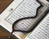 Ein Koran mit Perlenkette liegt auf einem Holztisch. Die Seiten sind mit arabischer Schrift bedruckt, die an die Geschichte von Yajuj und Majuj im Islam erinnert.