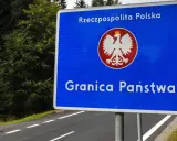 Niebieski znak z orłem i napisem "Rzeczpospolita Polska" oraz "Granica Państwa" stoi przy drodze otoczonej lasem.