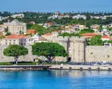 Widok na zabytkowe mury obronne i zabudowania wyspy Rodos, z błękitnym morzem w tle.