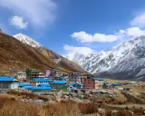 Villaggio colorato ai piedi dell'alta montagna himalayana, con case dai tetti blu e rosa, circondato da pendii bruni e cime innevate.