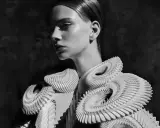 A model showcases intricate, sculptural white garments, embodying futuristic fashion. The avant-garde design features layered, organic shapes that create a dramatic silhouette.