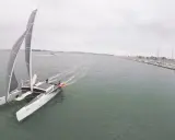 Un catamaran TS3 navigue sur une mer calme, avec un port rempli de voiliers à droite.