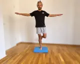 Mann trainiert den sprunggelenk auf einer blauen Matte, steht auf einem Bein, Arme ausgestreckt.
