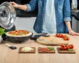 Otwarta opiekacz do pizzy, na kamieniu piec pizza z pomidorkami i bazylią. Obok deska z pomidorkami, startym serem i szynką.