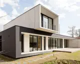Une maison moderne aux lignes épurées, avec des blocs de béton et de crépi sombre, dotée de grandes baies vitrées et d'une terrasse en bois.