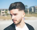Młody mężczyzna z modną fryzurą dla mężczyzn, z wygolonymi bokami i dłuższymi włosami na górze, na tle plaży.