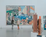 Two people observe a large, colorful mural depicting a chaotic scene with a blue car, factories, and people, offering a glimpse into the art market definition of diverse artistic expression.