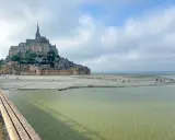 St. Michel, eine beeindruckende Abtei auf einer Felseninsel, ragt über einer sandigen Bucht auf. Menschen spazieren am Strand, während das Wasser sanft ansteigt.