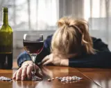 Kobieta z głową opartą na rękach, obok kieliszek wina, butelka i opakowania psychotropów.