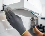 Hände mit Handschuhen ziehen einen Filter aus einer Dunstabzugshaube, um den Motor zu reinigen.