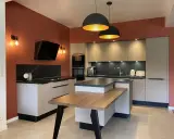 Cuisine moderne aux murs terracotta, avec îlot central en bois et mobilier gris clair. Éclairage chaleureux.