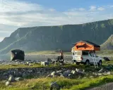 En man tar en selfie vid sitt tak-tält på en strand i Norge. Bredvid står en mörk kupoltält och ett bord med stolar. Berg i bakgrunden.