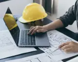 Inżynier analizuje plany architektoniczne, projektując powierzchnię wewnętrzną budynku. Na biurku leży żółty kask.