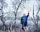 Britta Marakatt Labba i samedräkt håller upp en stor vit fjäder i en snötäckt skog.