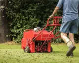 Man pushing a red core aeration machine across a green lawn, leaving plugs of soil behind.