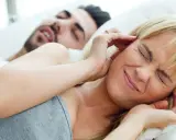Frau hält sich die Ohren zu, genervt vom Schnarchen ihres Partners, der trotz Seitenlage laut schnarcht.