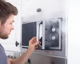 Mann öffnet Mikrowelle, aus der Rauch dringt. Ein Fehler mit Metall in Mikrowelle kann gefährlich sein.