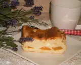 Pyszny sernik z brzoskwiniami, udekorowany gałązką lawendy, obok białej filiżanki i czerwono-białej łyżeczki.