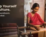 A woman in a red sari weaves on a loom, showcasing the rich tradition of handlooms of India. Stacked fabrics are visible.