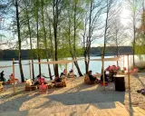 Pola namiotowe nad jeziorem, ludzie odpoczywają na plaży pod baldachimami, wśród drzew.