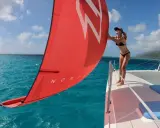Une femme en bikini ajuste la voile rouge d'un petit catamaran habitable. L'eau turquoise et le ciel bleu créent un décor paradisiaque.