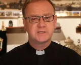 Wojciech Węgrzyniak w okularach, w czarnej sutannie z białą kolaretką, przemawia. W tle widać zwoje Tory i obrazy.
