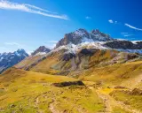 Ampia **foto grandangolo** di montagne innevate e pendii dorati autunnali, con un sentiero che serpeggia tra l'erba.