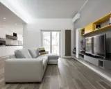 Un progetto soggiorno stretto e lungo con divano grigio, TV a parete e mensole gialle. La cucina moderna è visibile sullo sfondo.