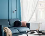Divano blu petrolio con cuscini decorativi e coperta. Parete azzurra con applique. Abbinamenti moderni e rilassanti.
