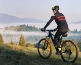 Rowerzysta w kasku i sportowym stroju pokonuje górski szlak, utrzymując dynamiczną kadencję rower. Mgła spowija doliny, tworząc malowniczy krajobraz.