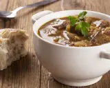 Aromaticzny gulasz wołowy w białej miseczce, ozdobiony natką pietruszki, obok kromka chleba i łyżka.