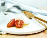 Kuleczki mozzarelli z pomidorkami koktajlowymi na białym talerzu, obok złote sztućce i ściereczka.