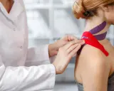 Fizjoterapeuta aplikuje czerwone i fioletowe tejpy na ramieniu i szyi pacjentki. Czy tejpy mogą zaszkodzić? Zastosowanie ich wymaga wiedzy.