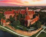 Zamek krzyżacki w Malborku, imponująca twierdza z czerwonej cegły, otoczona fosą i zielenią, pod rozgwieżdżonym, różowym niebem. Ciekawostki o jego historii czekają na odkrycie.