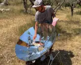 Un homme cuisine avec un four solaire parabolique, démontrant le fonctionnement de cet appareil écologique en plein air.